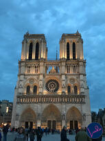 The illuminated towers and rose window of Notre-Dame Cathedral in Paris at dusk, with people gathered in the square.