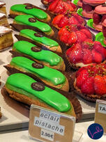Bright green pistachio éclairs displayed in a Paris pastry shop case, alongside strawberry tarts and macarons.